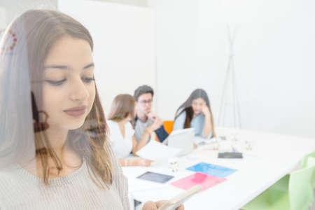 Shot through glass of young woman with smartphone and students on background.