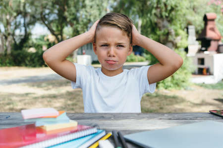 Little schoolboy looking away while sitting at table in garden with textbooks and school supplies.
