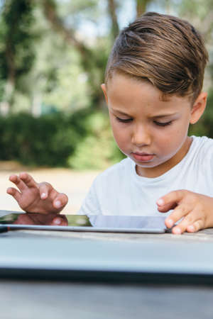 Portrait of little concentrated schoolboy sitting with tablet at table in summer garden.の写真素材