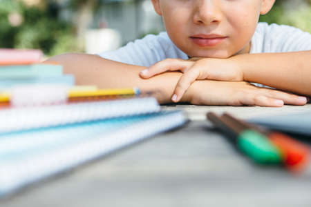 Crop shot of kid leaning head on hands while posing at table with notepads and pens.