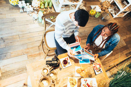 View from above of young black race woman and caucasian young man photographers co-working while having breakfast at agencyの写真素材