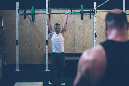 Young man doing jerks with barbell in front of his colleague on a fitness routine at the box gymの写真素材