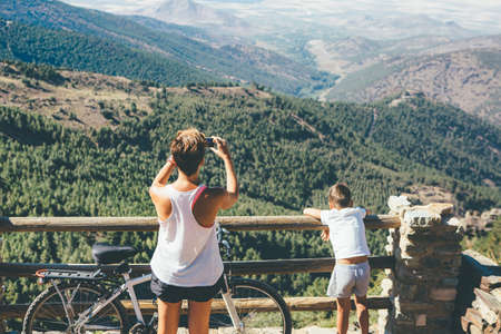 Close up of a back view of a girl taking picture with a smart phone of the landscape with a child resting after a bicycle ride. Copy space area availableの写真素材
