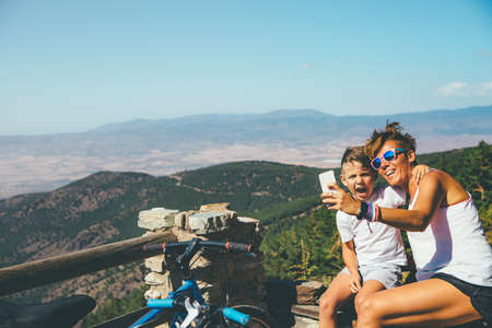 Happy young woman and child with bicycle taking a self photo with the mobile phone at the top of a mountain on a sunny day. Copy space area availableの写真素材