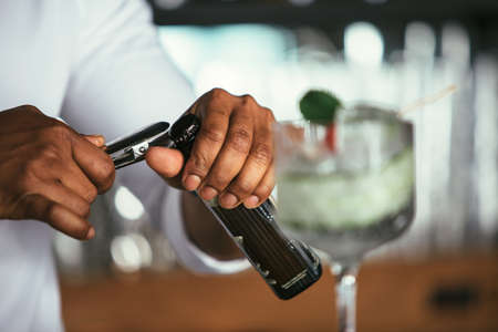Close up of the detail of the hands of a mixed race male expert bartender opening a bottle to...