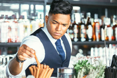 Mixed race male expert bartender is taking a cinnamon stick for decorating a cocktail at the bar counterの写真素材