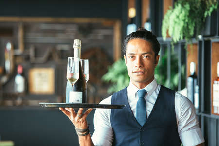 Close up of an elegant latin waiter is holding a tray with two glasses and a bottle of champagne in...