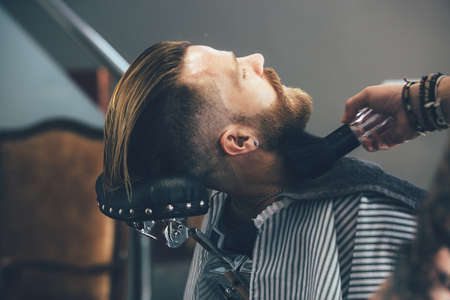 Hipster bearded man sitting in a chair is getting a beard cut in a barber shopの写真素材