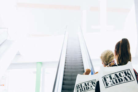 Black Friday. Back view of couple of young women shoppers holding paper shopping bags near an...