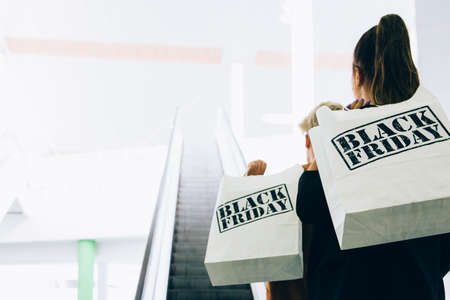 Black Friday. Back view of couple of female shoppers holding paper shopping bags near an escalator in a mall. Indoor shoot. Copy space area available.の写真素材