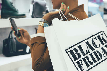 Black Friday. Close up of a cropped view of a young woman holding paper shopping bags while is looking to a mobile phone in a shoe shop.の写真素材
