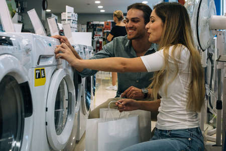 Side view of a smiling young couple choosing a washing machine in a home appliance store. Cyber...