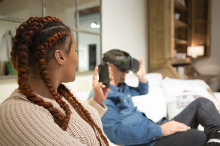 Young man is using virtual reality glasses while his young friend is using a smart phone making a...