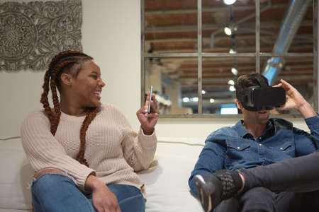 Happy young man using virtual reality glasses while his young friend using technology are having fun sit in the sofa at home.の写真素材