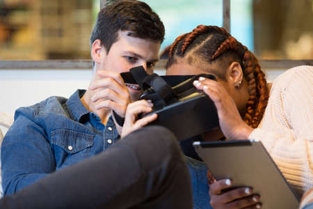 View of a young couple of friends, a caucasian man and a mixed race woman using virtual reality glasses and a tablet sitting in a sofa at home.の写真素材