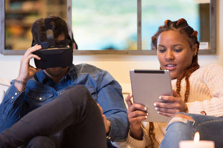 Couple of friends, a young caucasian man is using virtual reality glasses and a young mixed race woman is using a tablet sitting in a sofa at home.の写真素材