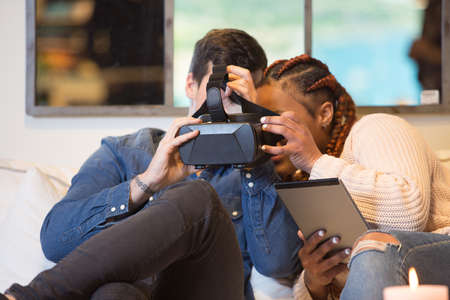 Young couple of friends, a caucasian man and a mixed race woman using virtual reality glasses and a tablet sitting in a sofa at home.の写真素材
