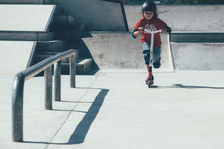 Front view of a young boy wearing a helmet and the protections is using a scooter in an urban skate park.の写真素材