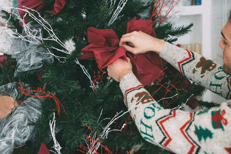 Cropped view of a young man and a young woman are decorating a fir tree with Christmas ornaments wearing Christmas sweatersの写真素材