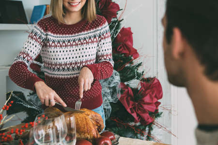 Cropped view of a young woman standing carves a roasted turkey on a Christmas table decorated in front of her beloved manの写真素材