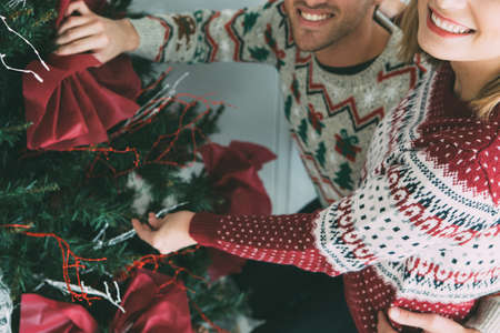 Cropped view of a smiling young woman and a young man decorate a fir tree with Christmas ornaments wearing Christmas sweatersの写真素材