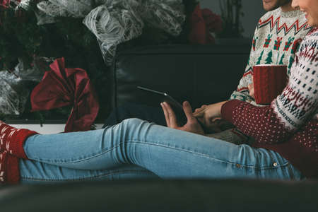 Cropped view of young woman and man are enjoying Christmas time using a tablet wearing Christmas sweaters sitting on a sofa at homeの写真素材