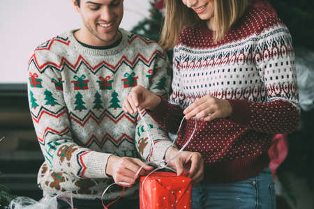 Cropped view of smiling young man and woman wrap a Christmas present wearing Christmas sweaters at homeの写真素材
