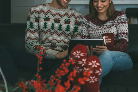 Cropped view of a lovely smiling young woman and man are enjoying Christmas time using a tablet wearing Christmas sweaters sitting on a sofa at homeの写真素材