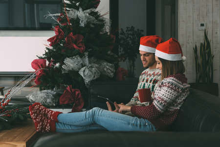 Lovely young man and woman are enjoying Christmas time using technology wearing Christmas sweaters at homeの写真素材