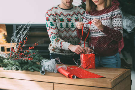 Cropped view of young woman and man are wrapping a Christmas gift wearing Christmas sweaters at homeの写真素材