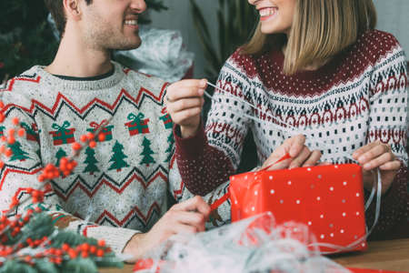 Cropped view of smiling young man and woman wrap a Christmas present box wearing Christmas sweaters at homeの写真素材