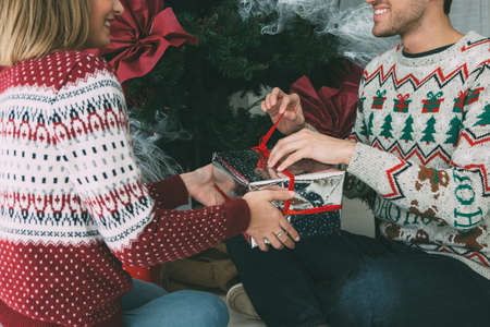 Cropped view of a smiling young man is giving a Christmas gift to his smiling beloved woman under the fir tree and wearing Christmas sweatersの写真素材
