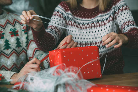 Cropped view of smiling young man and woman wrap a Christmas gift wearing Christmas sweaters at homeの写真素材