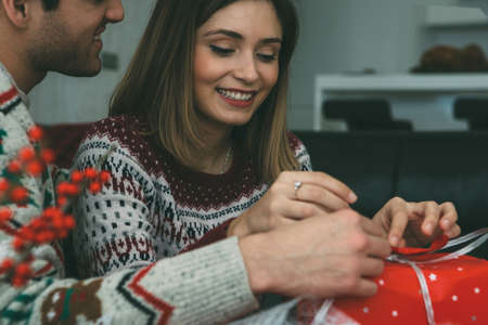 Smiling young woman and man are wrapping a Christmas gift wearing Christmas sweaters at homeの写真素材