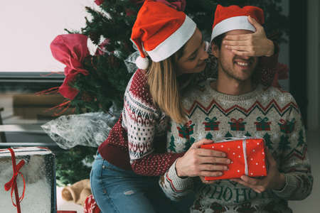 Smiling young woman is giving a Christmas gift to her beloved partner under the fir tree and wearing...