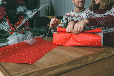 Cropped view of smiling young woman and man are wrapping a Christmas present box wearing Christmas sweaters at homeの写真素材