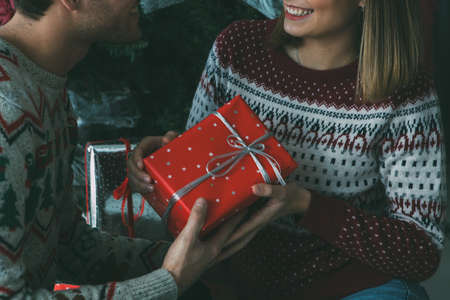 Cropped view of a smiling young woman is holding a Christmas gift with her beloved man under the fir tree and wearing Christmas sweatersの写真素材