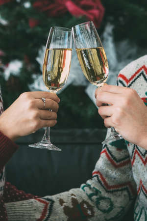 Cropped view of the hands of a young man and young woman are holding drinking glasses and toasting for Christmas near the fir tree and wearing Christmas sweatersの写真素材