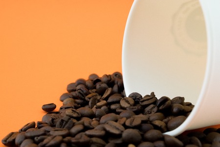 Paper coffee cup with spilled coffee beans, orange background. Roasted coffee beans.の写真素材