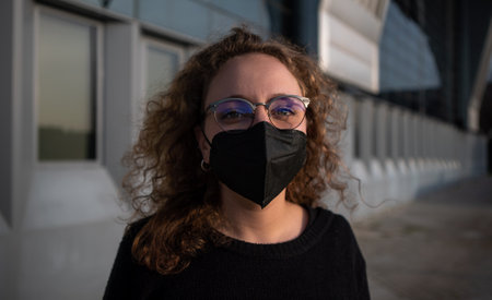 Young woman wearing a mask to avoid contagion of covid 19 near a building. The woman wears glasses and has curly blonde hair.の写真素材