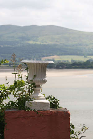 An elegant white urn stands on a plinth, entwined with plants. In the background is, just visible, a beautiful seascapeの写真素材