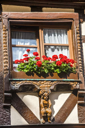 Ribeauville (Bas-Rhin, Alsace, France) - Exterior of old half-timbered house: window with wooden statue and red flowersの写真素材