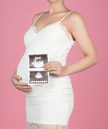 Pregnant woman holding ultrasound scan on her belly isolated on pink backgroundの写真素材
