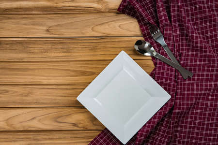 A white plate rests on checkered tablecloth. There are spoon and fork on wood backgroundの写真素材
