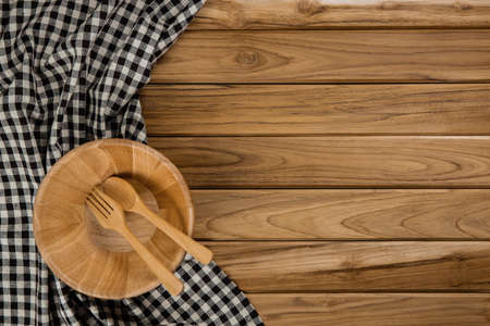 A wood bowl rests on checkered tablecloth. There are spoon and fork on wood background
