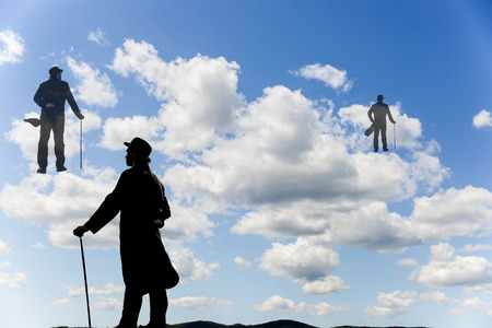 Surrealistic image of silhouettes of men with a cane and bowl hat climbing up the clouds in the skyの写真素材