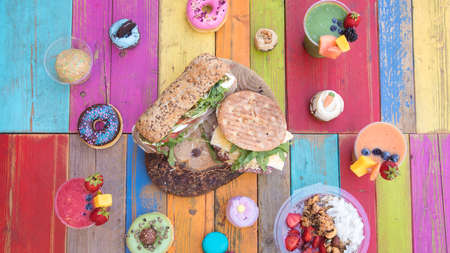 Colorful and tasty drinks and snacks with smoothies, cupcakes, donuts and sandwiches on a colorful wood tableの写真素材