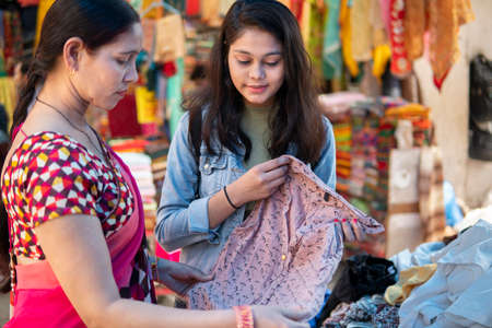 Happy mother and daughter looking and buying clothes together from outdoor street market of Delhi,...