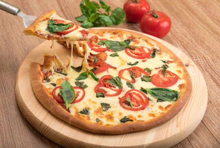 Pizza Margherita with tomatoes, mozzarella and basil on a wooden background, a slice of pizza with cheese stretching, close-upの写真素材