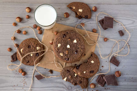 Chocolate cookies with hazelnuts, white chocolate and dark chocolate on parchment and glass of milk, wooden background, flat lay, top viewの写真素材